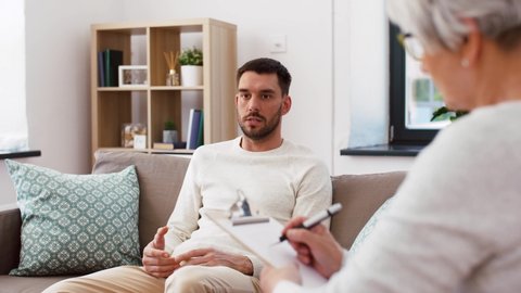 psychology, mental therapy and problem concept - senior woman psychologist listening to young man patient and taking notes on clipboard at psychotherapy session