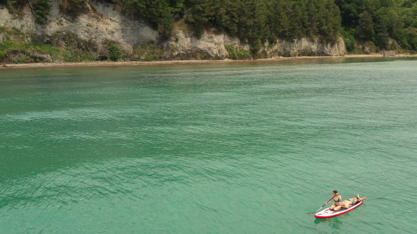 Sup surfer woman rowing on supboard. Paddle board sailing on turquoise water along beautiful green cliff. Beautiful seascape. Man resting lying on surfboard. Outdoor summer water sport aerial view