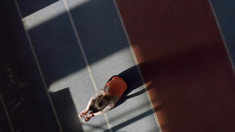 Pretty girl in an orange T-shirt with black pants and light sneakers is doing a stretching exercises at the indoor stadium. Sun is shining onto her body. Top view video recording.