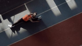 Cute girl in an orange T-shirt with black pants and light sneakers is doing a stretching exercises at the indoor stadium. Sun is shining onto her body. Top view video recording.