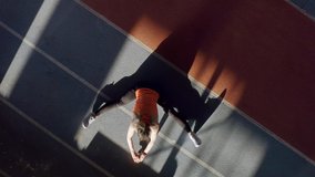 Athletic girl in an orange T-shirt with black pants and light sneakers is doing a stretching exercises at the indoor stadium. Sun is shining onto her body. Top view video recording.