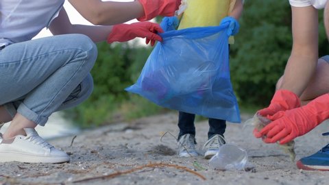 charity cleaning, young family with little kid in rubber gloves collects refuse in garbage bag on dirty beach from plastic while cleaning close-up on sand