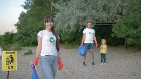 cleaning beach, portrait of young volunteer mother in rubber gloves with garbage bag near pointer sign on background father and little daughter after collecting plastic dregs on nature