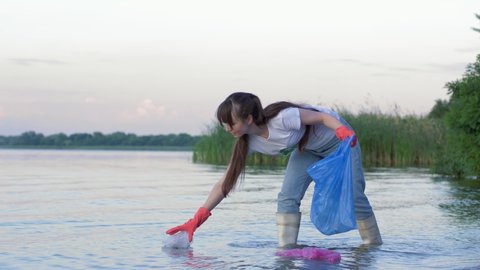 problem of plastic pollution, young volunteer woman into rubber boots cleans beach from refuse and collects polyethylene trash in garbage bag while standing in river water