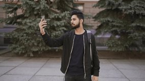 Young bearded indian man use phone for video call conversation while walking on the city street