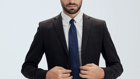 Cropped view of serious young businessman in black suit and blue tie buttoning his jacket and putting his hands in pockets over gray background isolated