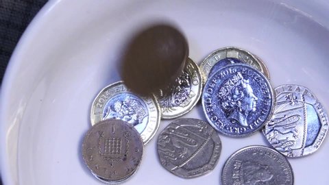 A paltry donation of small change in English UK currency is thrown into a white collection bowl. The money falls, rebounds and spins in slow motion.