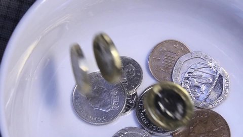 A  donation of assorted English UK coins are thrown into a white collection bowl in an act of charity. The money falls, rebounds and spins in slow motion.