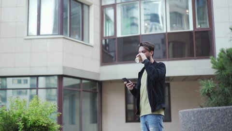 Young man receiving sms on his cell phone while walking down city street with takeaway coffee cup. Man stopping to read sms on phone, drinking coffee and proceeding walk texting back on the move