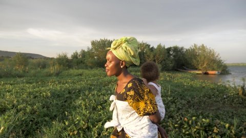A tracking shot from the side of an African mother with her baby on her back walking by a lake in the golden sunset light.