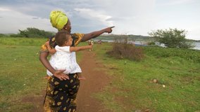 A tracking shot from behind of an African mother walking on a dirt track with her baby tied on her back near lake Victoria and past cows.