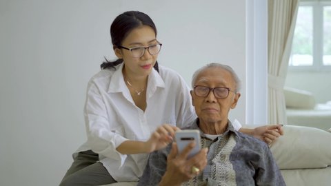 Young woman teaching elderly man how to use a smartphone on the sofa in living room at home. Shot in 4k resolution