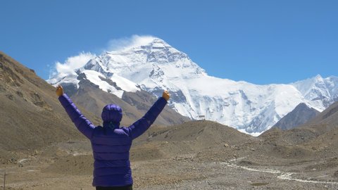 SLOW MOTION, CLOSE UP, COPY SPACE: Female trekker victoriously outstretches arms while observing snow capped Mount Everest. Excited woman on climbing adventure views windswept Everest from base camp.