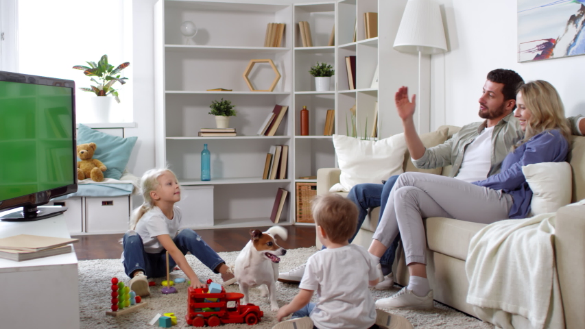 Medium shot of man sitting on sofa and talking while watching TV with cheerful young woman. Little children and Jack Russell terrier dog playing on living room floor Royalty-Free Stock Footage #1033907981