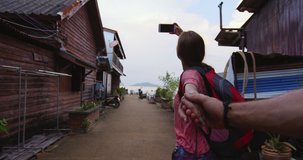 Young couple holding hands, tourist woman leading boyfriend, walking down alleyway in Ko lanta old town, taking photo or video with Smartphone. POV slow motion travel concept, hand held.