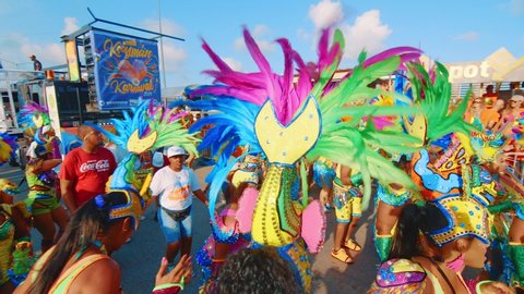 Willemstad, Curaçao - 03 03 2019: A crowd of people in vibrant neon costumes with elaborate headdresses dance and march down the street in the Carnival Gran Marcha in Curacao. Slow Motion High Angle P
