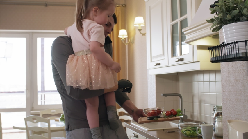 Tilting medium shot of handicapped Caucasian man holding up 3-year-old daughter with bionic prosthetic arm, making vegetable salad for lunch and talking on mobile phone pressed against shoulder