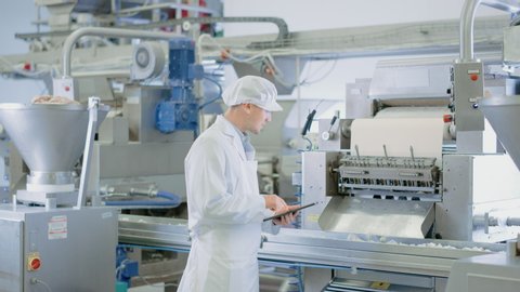 Young Male Quality Supervisor or Food Technician is Inspecting the Automated Production at a Dumpling Food Factory. Employee Uses a Tablet Computer for Work. He Wears a Sanitary Hat and Work Robe.