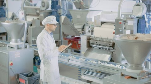 Young Male Quality Supervisor or Food Technician is Inspecting the Automated Production at a Dumpling Food Factory. Employee Uses a Tablet Computer for Work. He Wears a Sanitary Hat and Work Robe.
