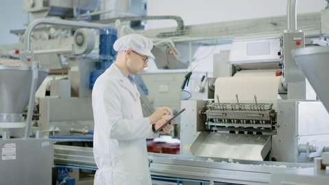 Young Male Quality Supervisor or Food Technician is Inspecting the Automated Production at a Dumpling Food Factory. Employee Uses a Tablet Computer for Work. He Wears a Sanitary Hat and Work Jacket.