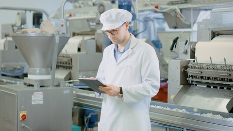 Young Male Quality Supervisor or Technician in Glasses is Working on a Food Factory. Employee Looks and Smiles at Camera. Expresses Success and Happiness. He Wears a White Sanitary Hat and Work Robe.
