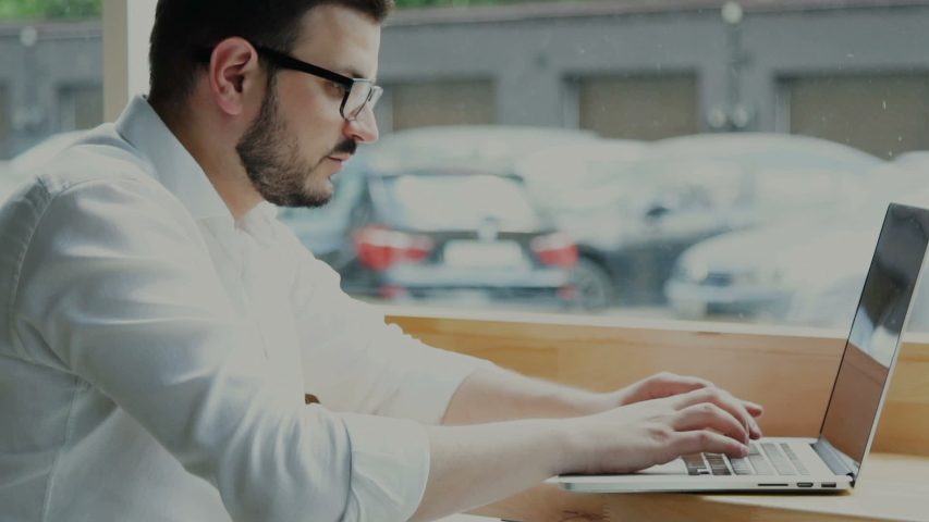 handsome businessman texting on laptop and using phone near window indoor, freelancer in formal clothes working in cafe