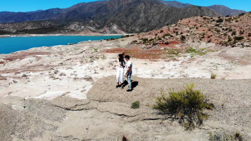 Aerial 4k drone shot orbit around a couple, on the coast, of the Potrerillos turquoise lake, on a sunny day, with mountain background in Mendoza, Argentina. Couple exploring Potrerillos