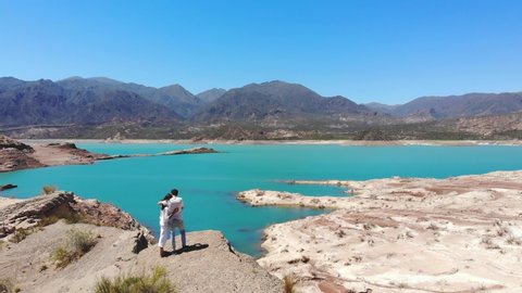 Aerial, drone shot, over a couple, looking at the blue water, of the Potrerillos lake, on a sunny day, in Mendoza, Argentina, South America