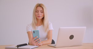 Closeup portrait of adult caucasian blonde businesswoman having a video call on the phone waving hi sitting in front of the laptop indoors in the office