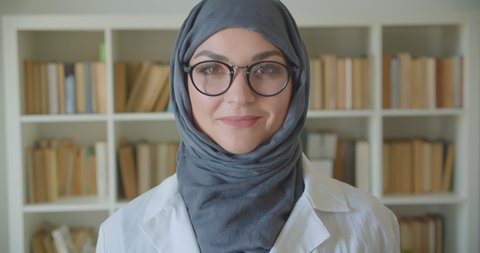 Closeup portrait of young muslim attractive female doctor in hijab and glasses looking at camera smiling happily standing in the library indoors