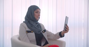 Closeup side view portrait of young muslim attractive female doctor in hijab and white coat having a video call on the tablet waving hi sitting in the armchair in the white room indoors