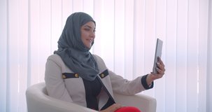 Closeup side view portrait of young muslim female doctor in hijab and white coat having a video call on the tablet sitting in the armchair in the white room indoors