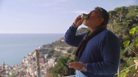 Elderly  Italian male retiree with an espresso cup looking out at the beautiful Amalfi Coast landscape with view of the ocean and skyline, with soft natural light. Wide shot on 8k helium RED camera