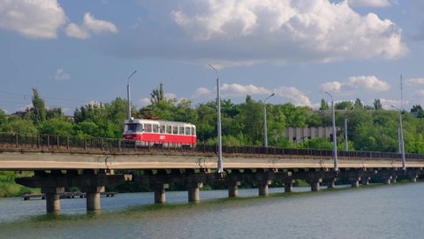 Tram moves along the bridge over the river in a city on a background of green trees and blue cloudy sky in Kryvyi Rih City, Ukraine.