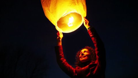 young woman holds above head glowing yellow hot chinese lantern at night, inside of it fire burns 