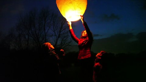 woman holds glowing yellow hot chinese lantern, son and daughter look, then she releases, and it flies up in night sky