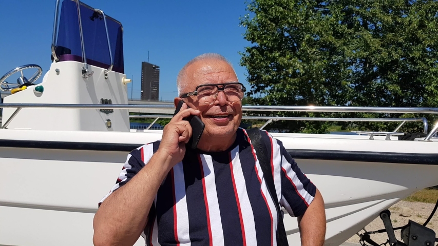 happy mature man in glasses stands with mobile phone at white cater on embankment against blue sky and highway closeup