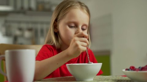 Close-up of a cute baby blonde girl in a red t-shirt with pleasure and appetite eating porridge at the table at home in the kitchen , morning healthy Breakfast
