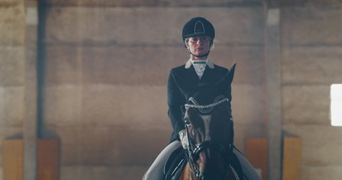 Cinematic slow motion close up of young female horsemanship master dressed in a professional apparel is practising exercises for competition of horse racing and dressage on a riding hall