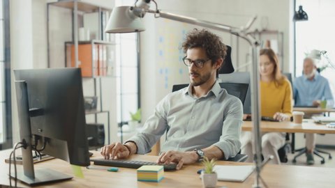 Handsome and Smart Hispanic Office Employee Sitting at His Desk Works on a Desktop Computer. In the Background Modern Bright Office with Diverse Group of Professionals Working for Growing Startup
