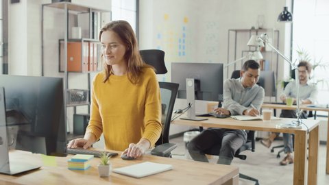 Beautiful and Smart Red Haired Female Specialist Sitting at Her Desk Works on a Desktop Computer. In the Background Modern Bright Office with Diverse Group of Professionals Working for Growing Startup