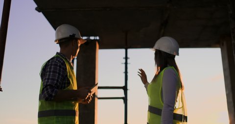 Builder man with a tablet and a woman inspector in white helmets shake hands at sunset standing on the roof of the building. Symbol of agreement of successful work.