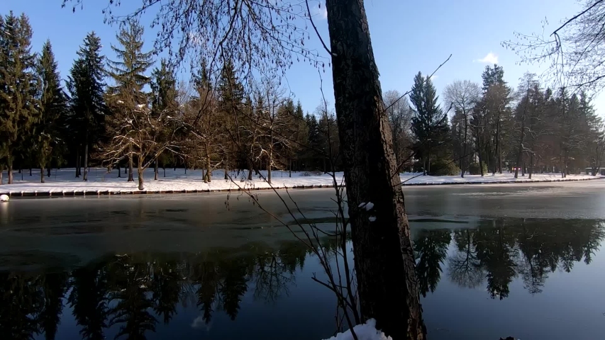 Slow left side pan of a park in winter, with the shore covered in snow and the lake half frozen.