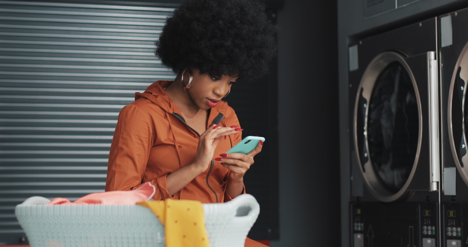 Smiling young woman using smartphone at laundromat. Self-service public laundry. | Shutterstock HD Video #1033997597