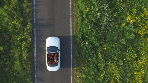 Aerial shot of white convertible car riding through empty rural road. Four young unrecognizable women travelling at cabriolet. Flying over auto driving at countryside road on summer day. Slow motion