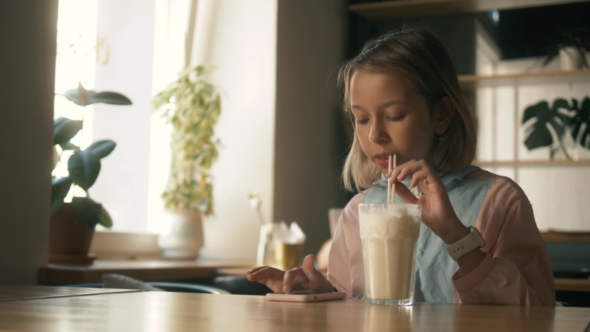 Beautiful girl in sitting in cafe counter and drinking milkshake while taking cute photos on her cellphone, child kid after lessons with cellphone | Shutterstock HD Video #1034002895