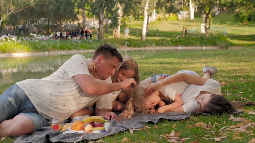 Family with kids having picnic outdoors. Parents with two children girls relax in a summer garden. Mother, father, little girl and baby boy eat fruit, drink juice for healthy lunch in a park.