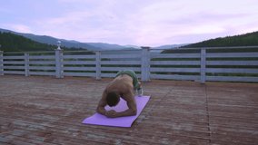 Muscular athlete with bare torso wearing green shorts and sport sneakers doing his abs on yoga mat. Strong man with athletic body training outdoors with amazing mountain view behind