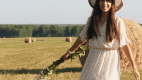 Woman in white dress and straw hat spinning round on green meadow with bouquet of daisies in arms and smiles broadly. Happy young woman at the summer meadow and enjoying freedom.