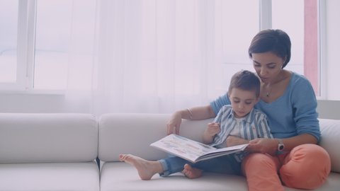 Happy mother and child son reading book laughing in bed. Happy family mother and child son reading holding book lying in bed, smiling mom baby sitter.